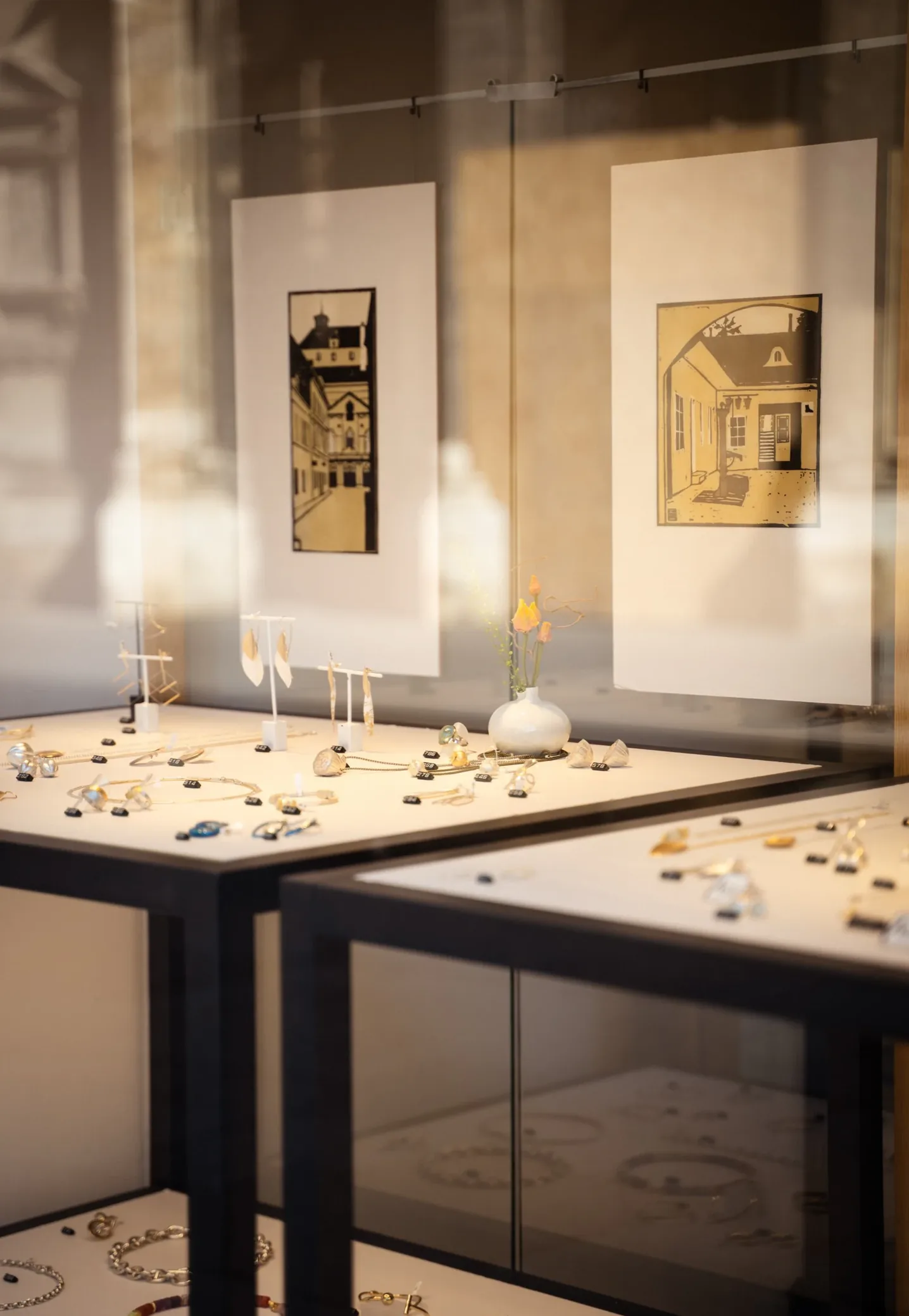 Glasvitrine mit Schmuck, dahinter gerahmte Grafiken an der Wand