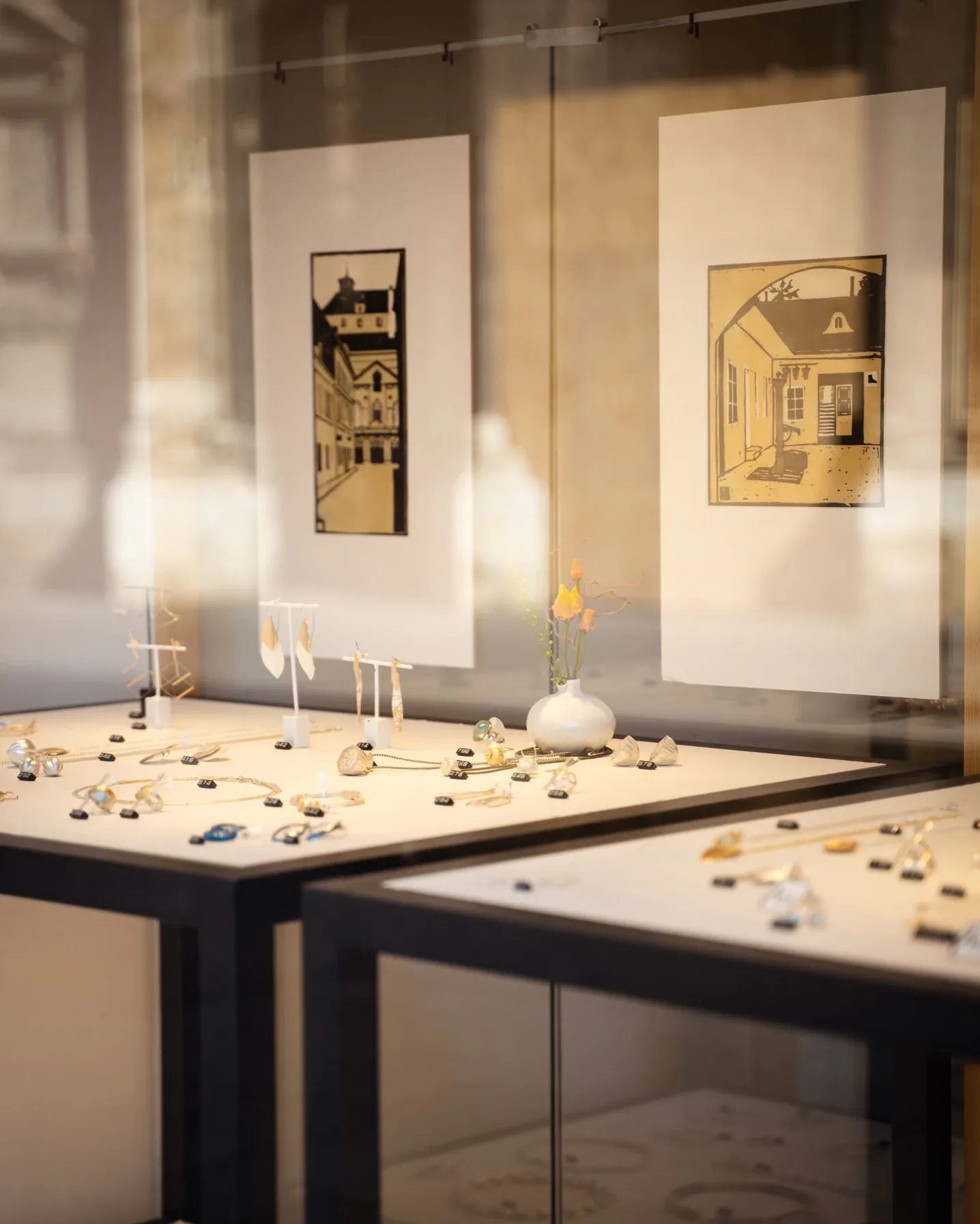 Glasvitrine mit Schmuck, dahinter gerahmte Grafiken an der Wand